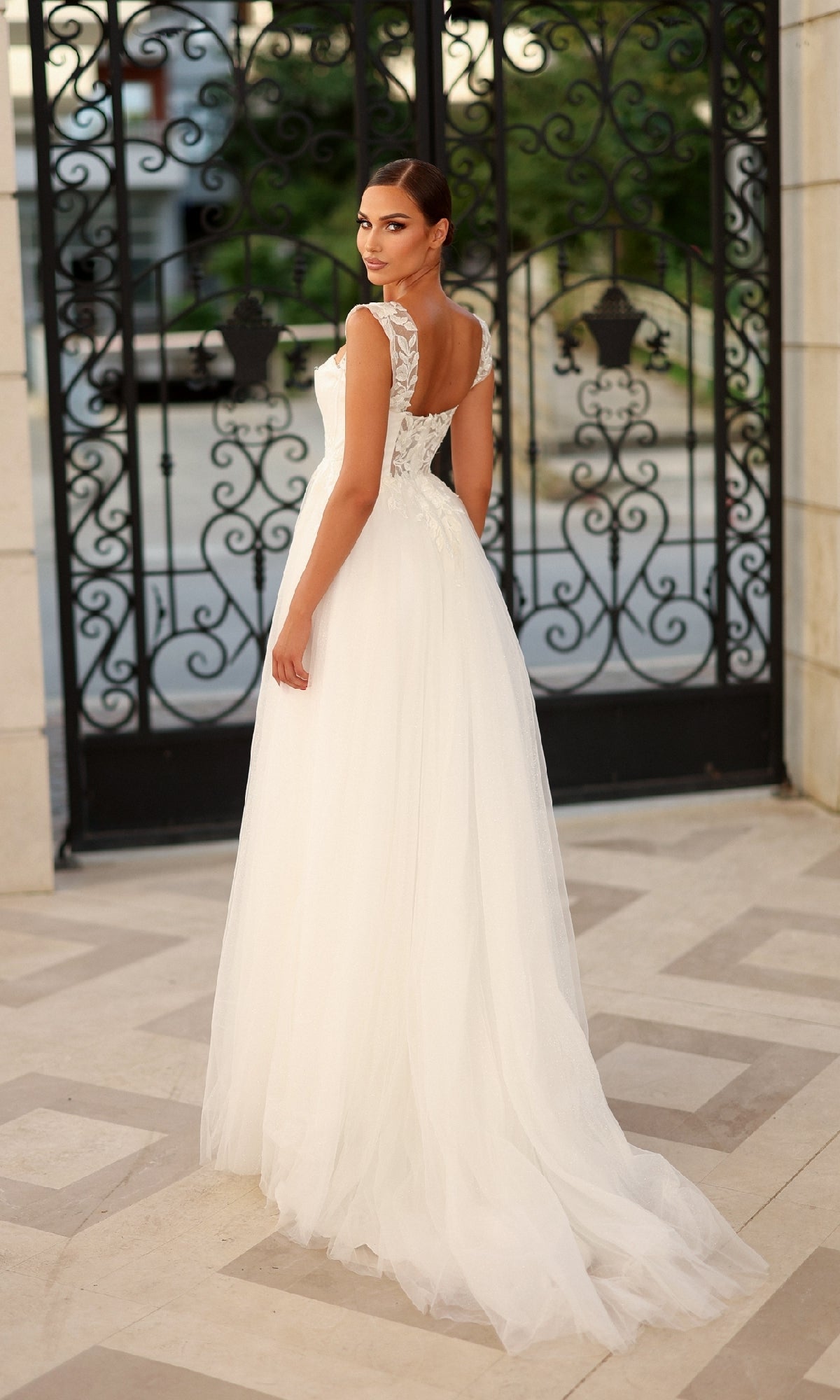 A woman wearing the Long Formal Dress: Tina Holly TW903, a white floor-length gown with lace appliques, stands outside in front of a decorative wrought-iron gate, looking over her shoulder.